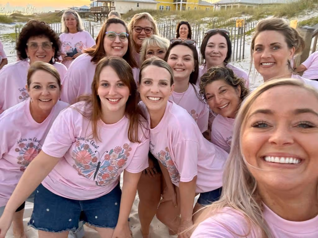 Group of women smiling and wearing matching pink T-shirts on a sandy beach at sunset.