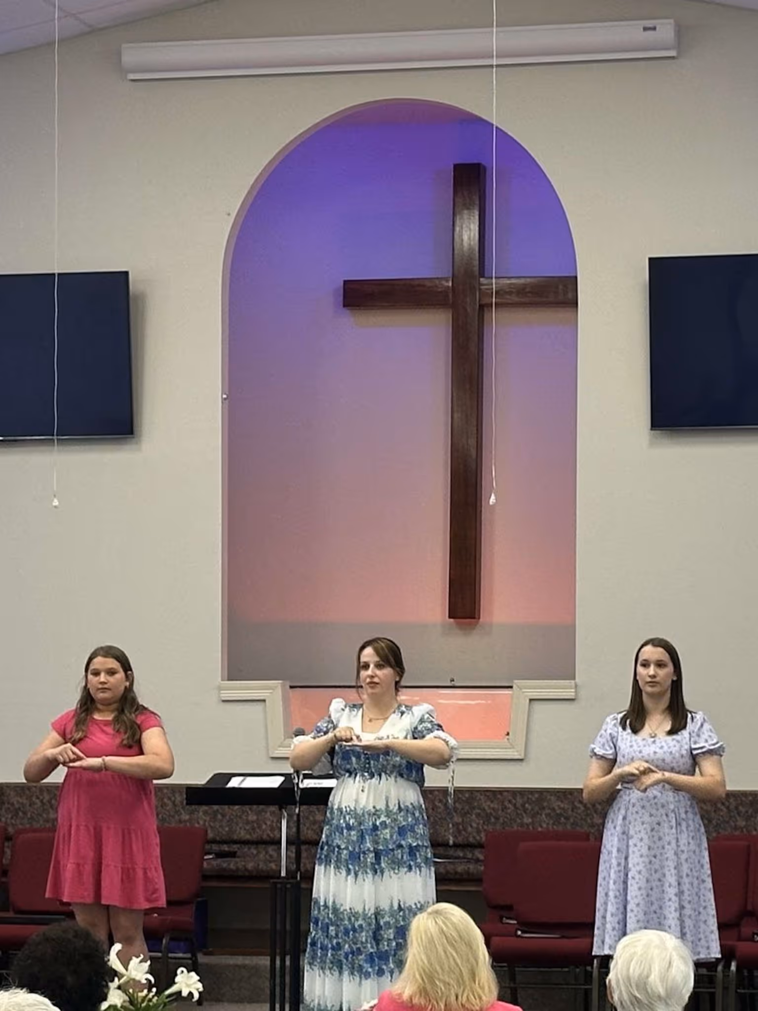 Three women standing in front of a cross in a church, performing sign language with hands.