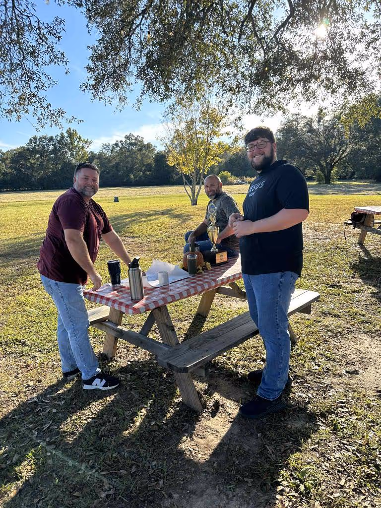 Three men standing and sitting around a picnic table outdoors with a trophy and drinks on it.
