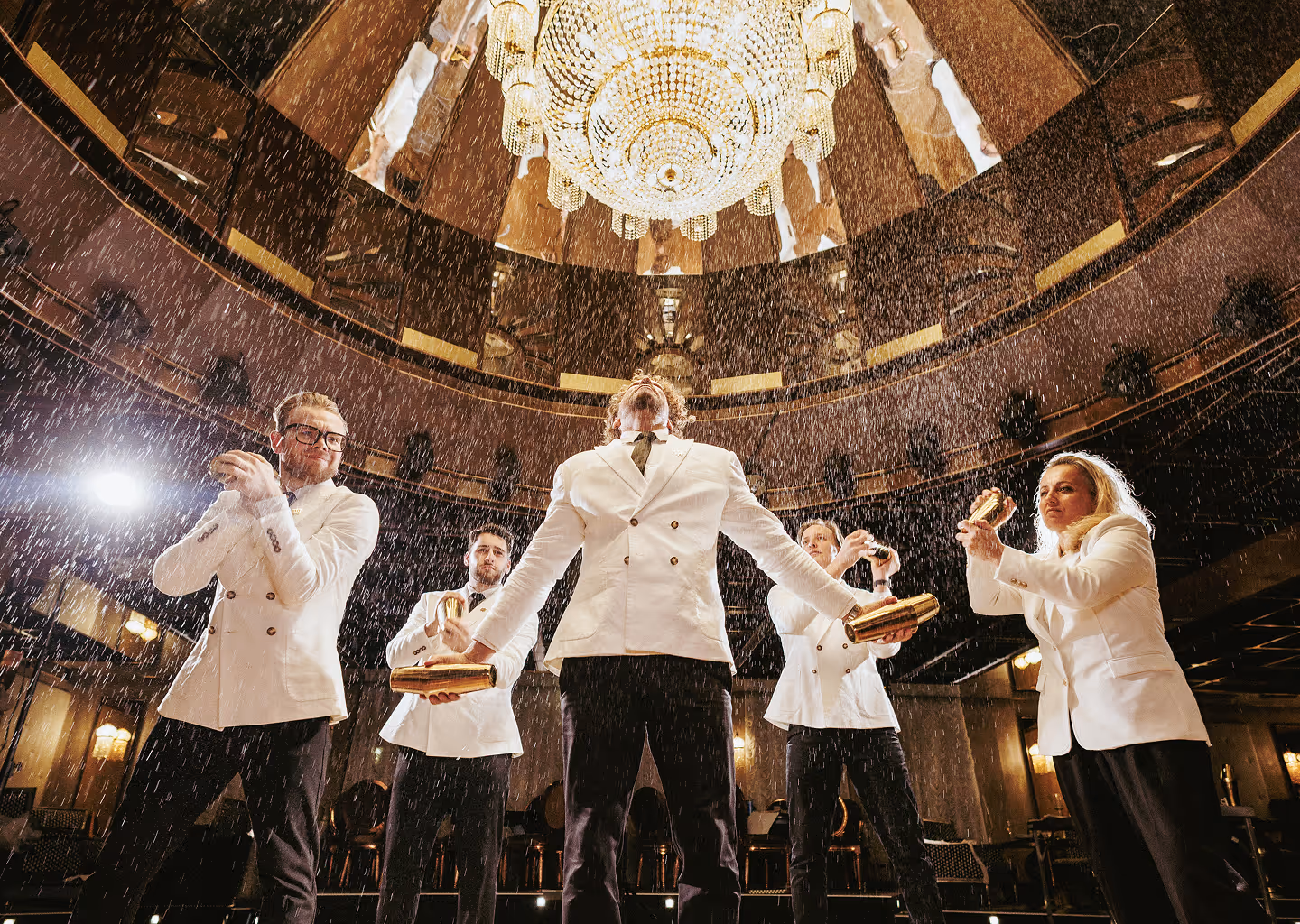 Five bartenders in white jackets shaking golden cocktail shakers beneath a large chandelier indoors.