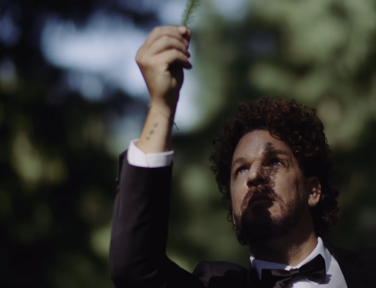 Mario Hofferer  in tuxedo holding a small green plant branch up outdoors with dappled light on his face.