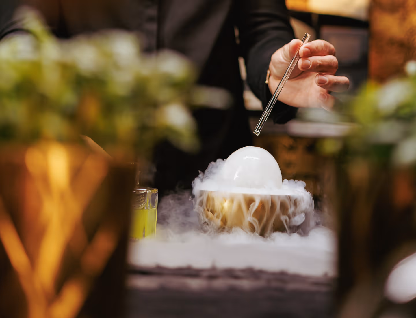 A hand holding a glass rod above a bowl with smoky vapor and a white spherical object inside.