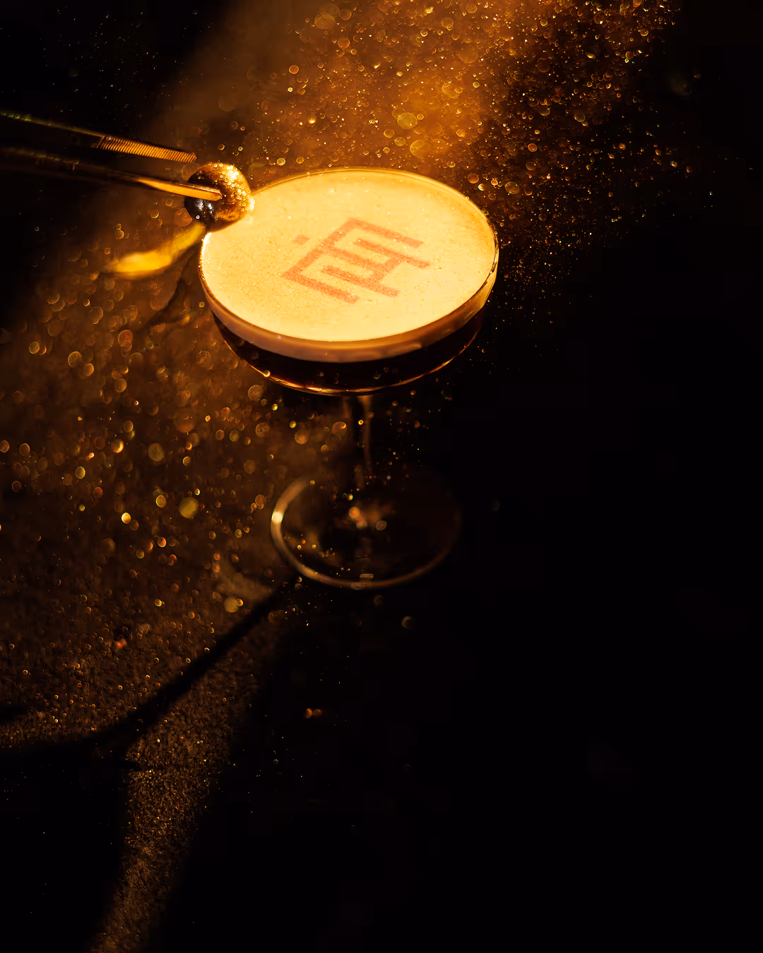 Cocktail glass with foamy drink and misty golden particles, a garnish being placed on top.