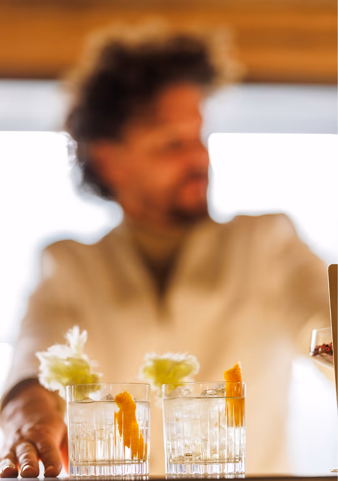 Two clear cocktails with ice and orange peel garnish in focus, person blurred in background.