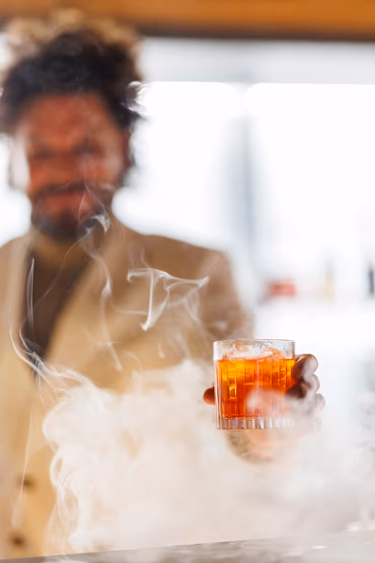 Man holding a glass of amber cocktail with ice surrounded by swirling smoke or mist.