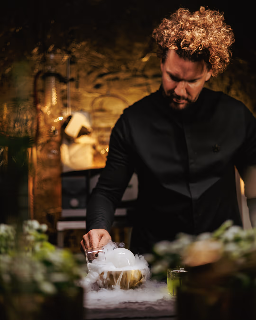 Mario Hofferer  with curly hair in black shirt pouring a smoking drink at a dimly lit table.