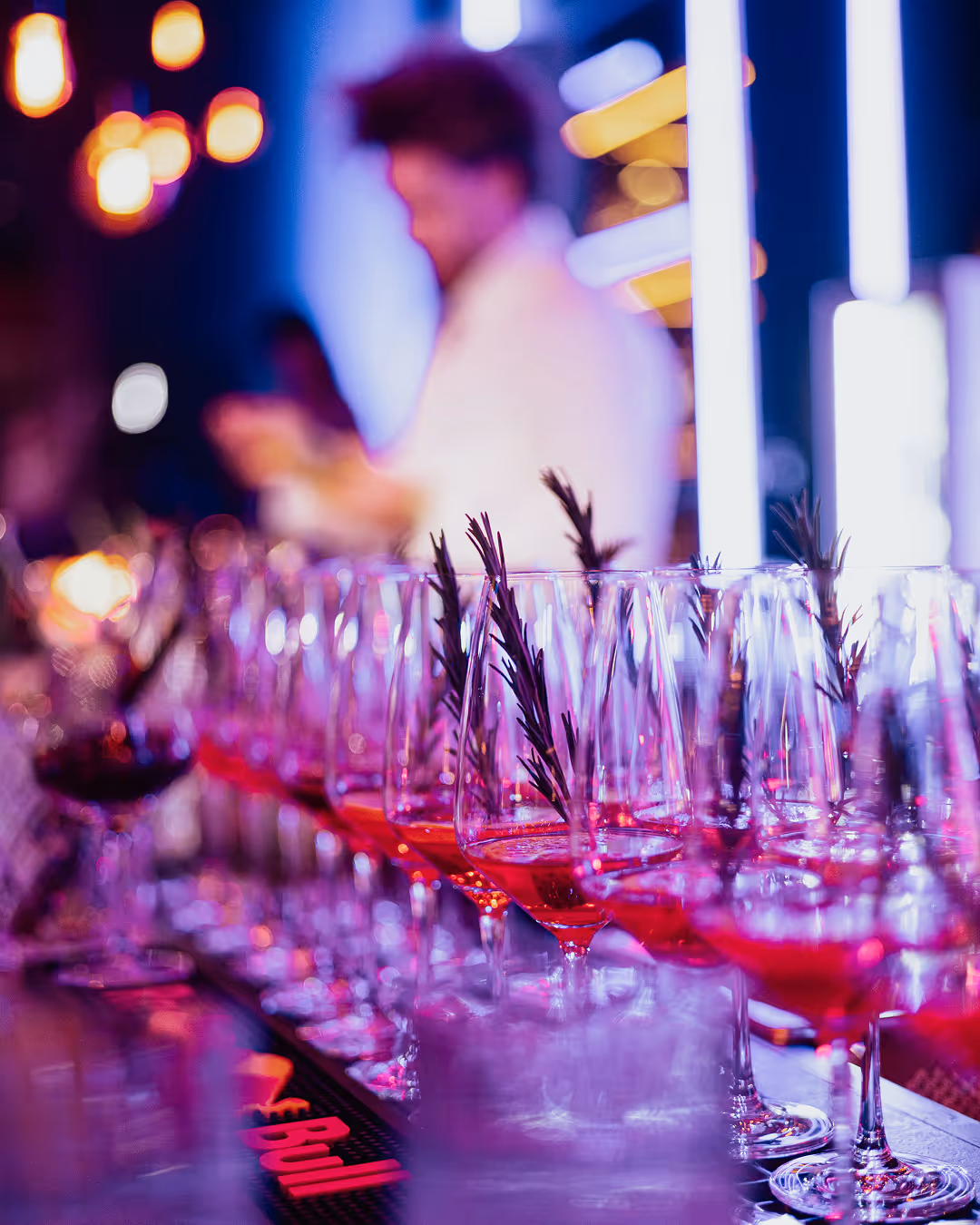 Row of cocktail glasses with red drink and rosemary garnish on a bar counter with blurred bartender.