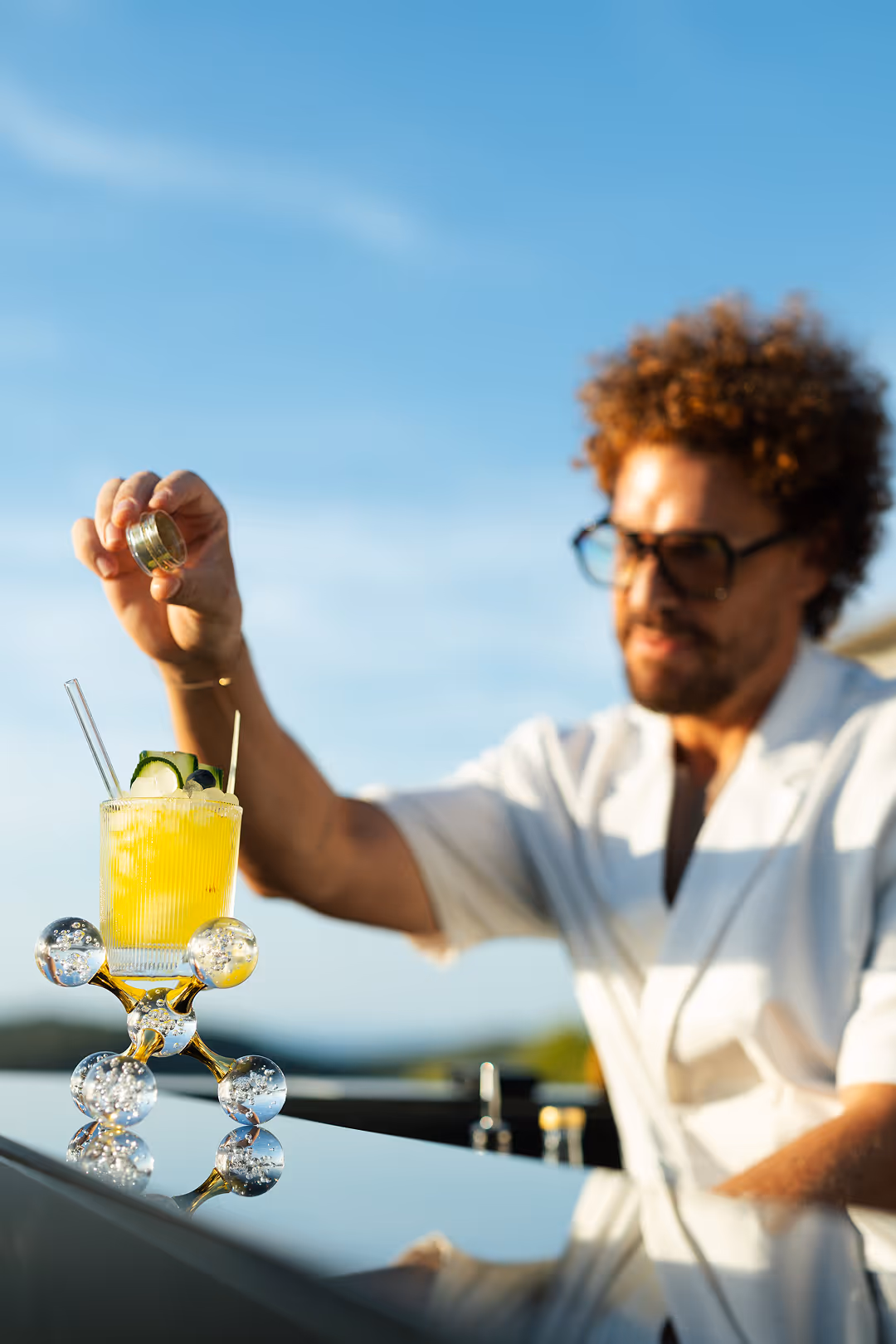 Man with curly hair and sunglasses preparing a yellow cocktail outdoors on a glossy bar surface.