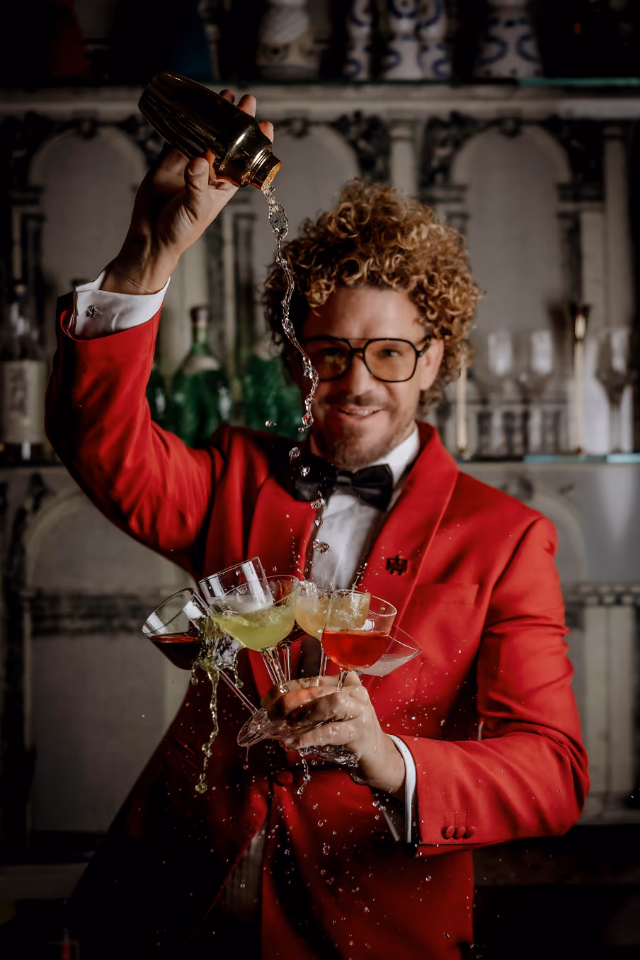 Bartender in red jacket pouring liquid from shaker into four cocktail glasses held in one hand.