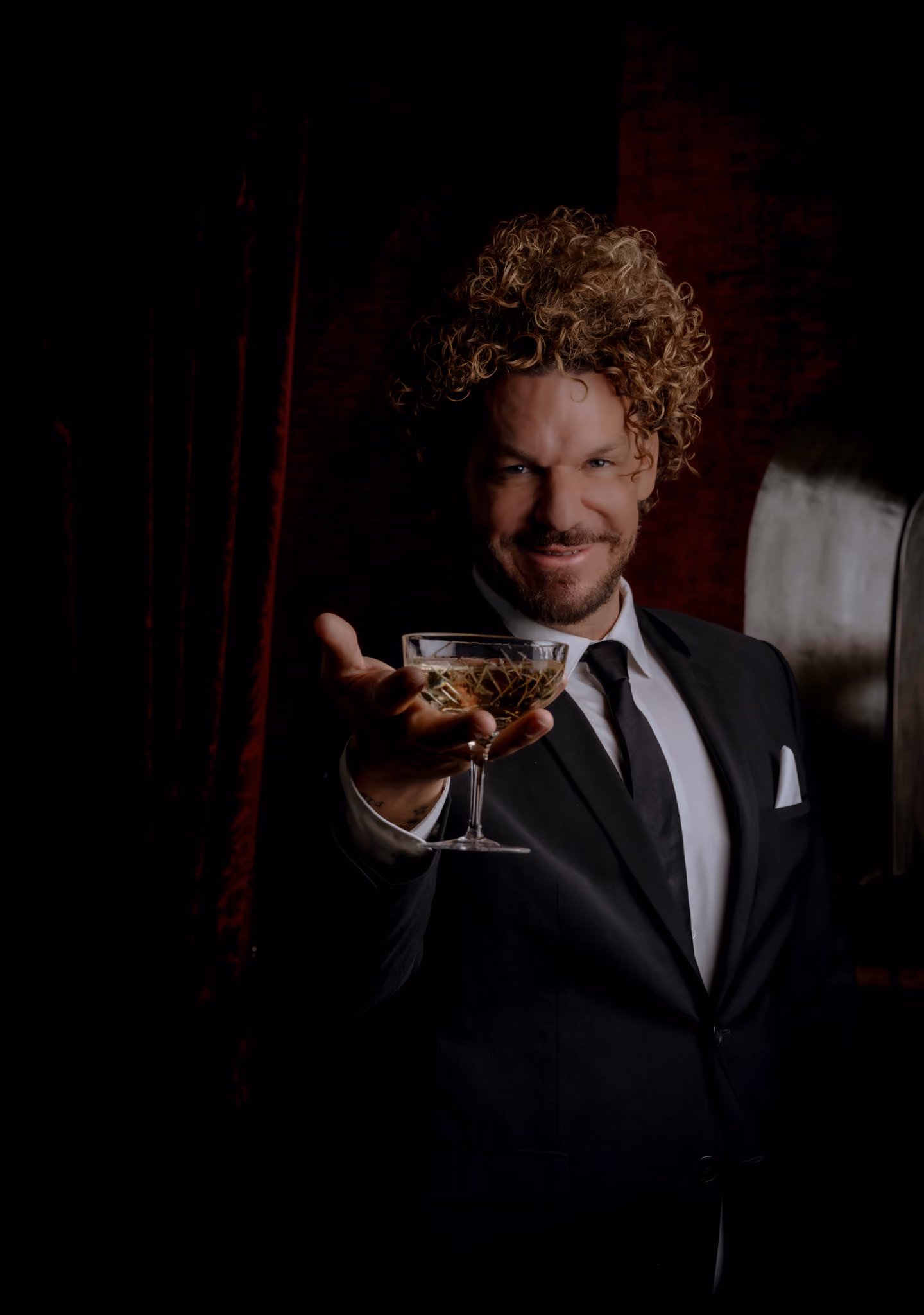 Man in white jacket pours red drink into a glass at a classical ornate indoor bar.