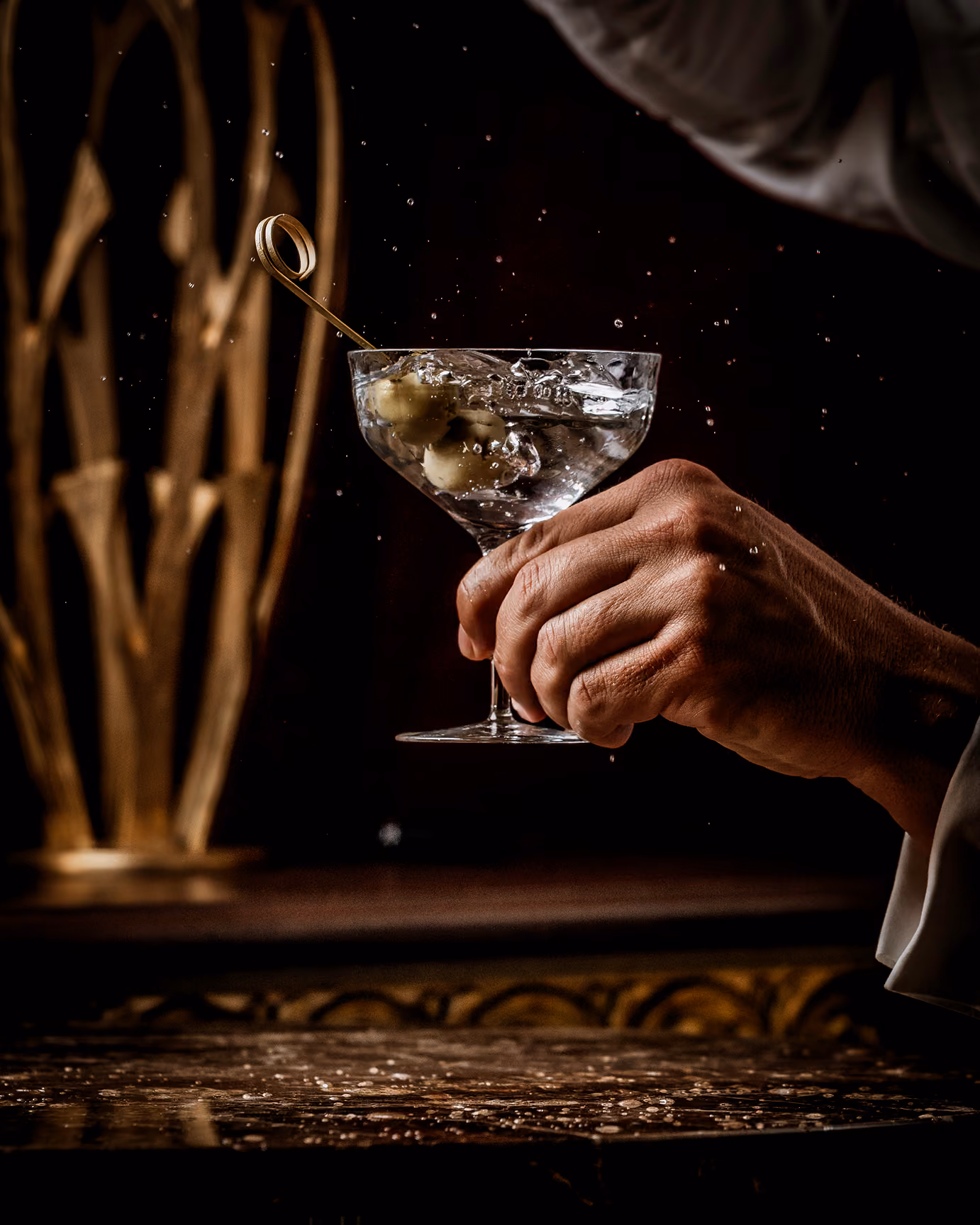 Hand holding a martini glass with olives and splashing liquid in a dimly lit setting.
