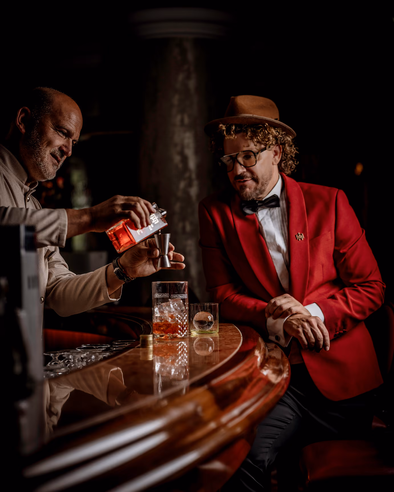 Mario Hofferer pouring liquor into a jigger while a man in a red jacket and hat watches at the bar.
