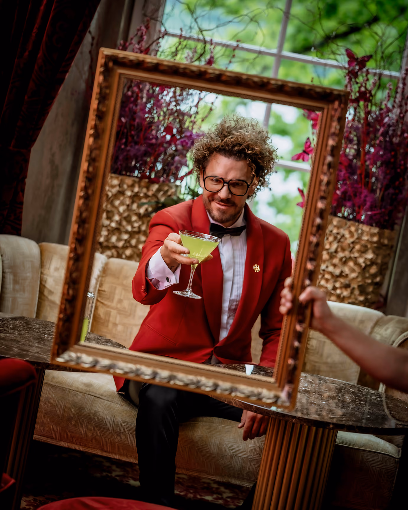 Mario Hofferer in a red jacket holding a green cocktail glass, framed by a large ornate picture frame.