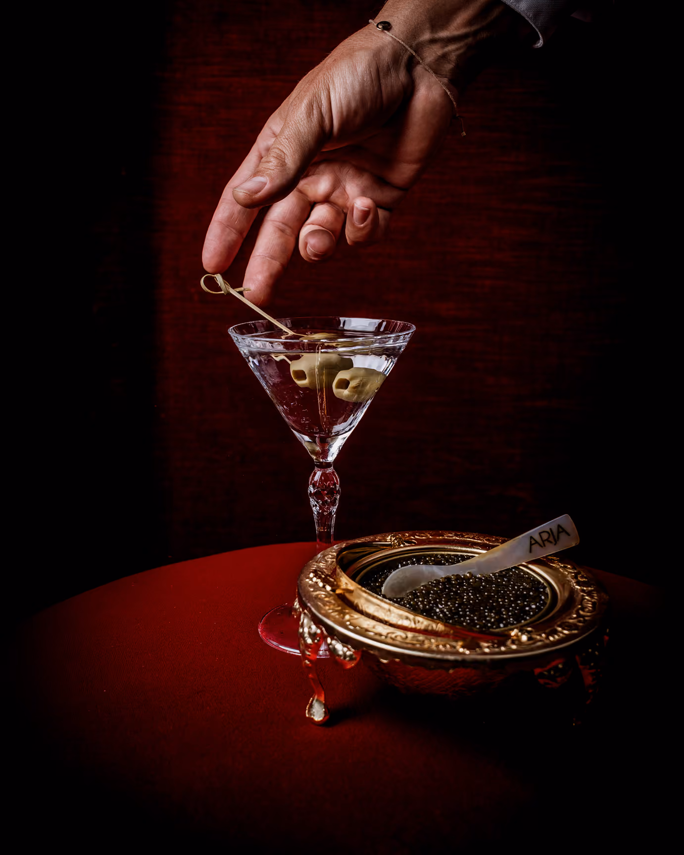 Hand holding a cocktail pick above a martini glass with olives, next to a gold bowl of caviar.