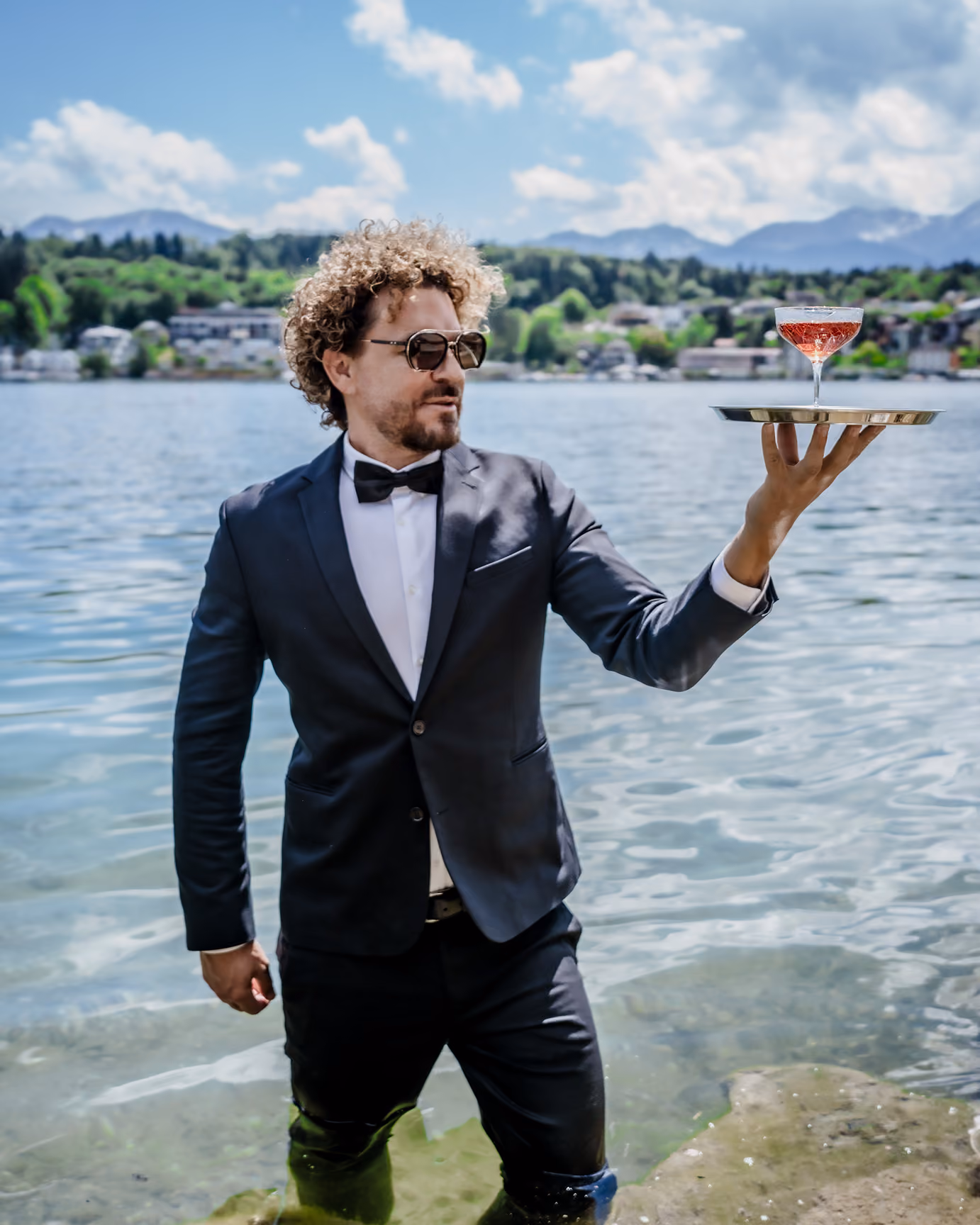 Mario Hofferer in suit with bow tie and sunglasses stands knee-deep in water holding a tray with a glass of red drink.