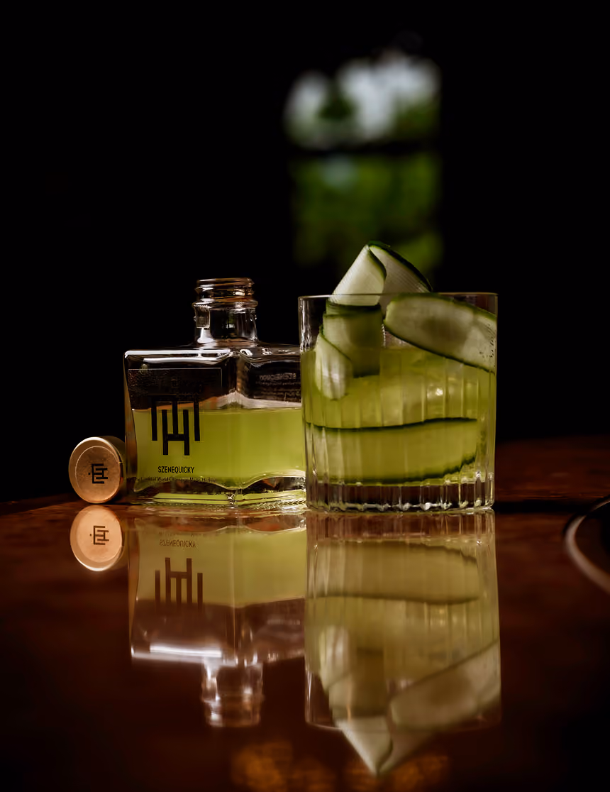 Glass of green cocktail with cucumber slices next to a partially filled bottle on a reflective surface.