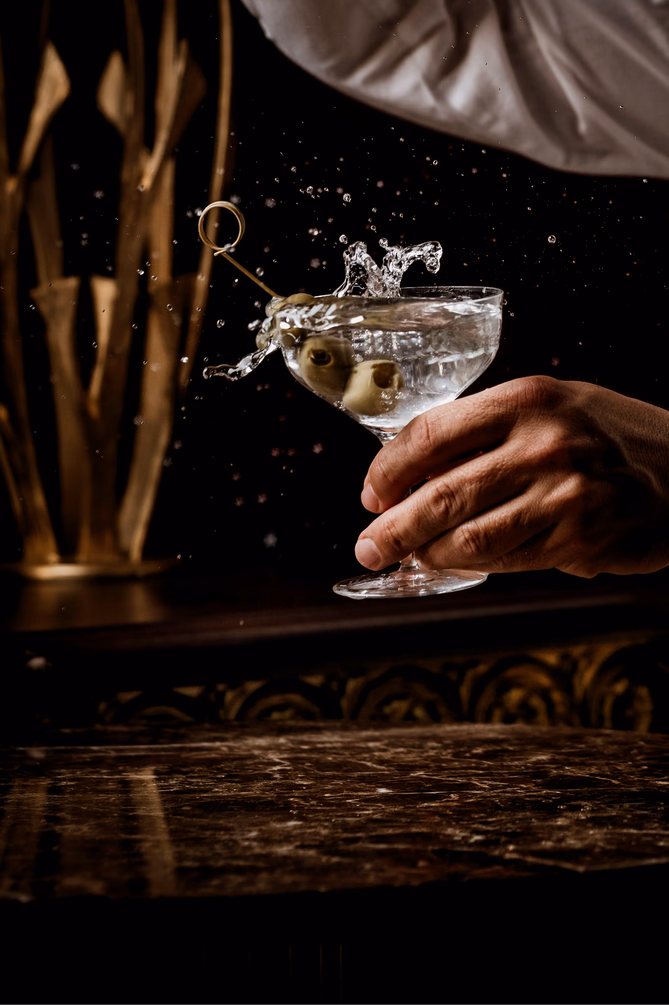 Hand holding a martini glass with olives and splashing liquid against a dark background.