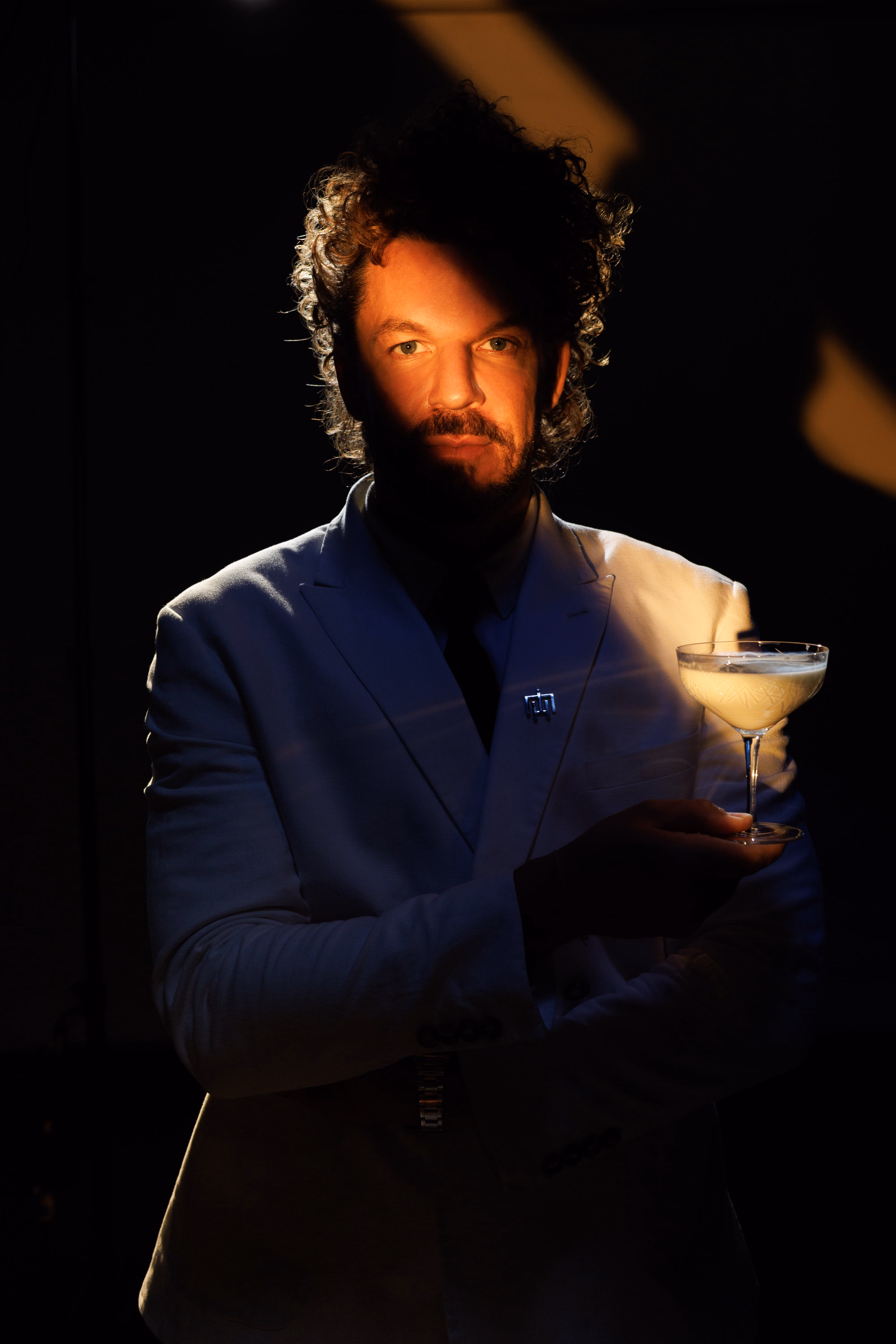 Mario Hofferer in white suit holding a cocktail glass in dim light highlighting his face and drink.