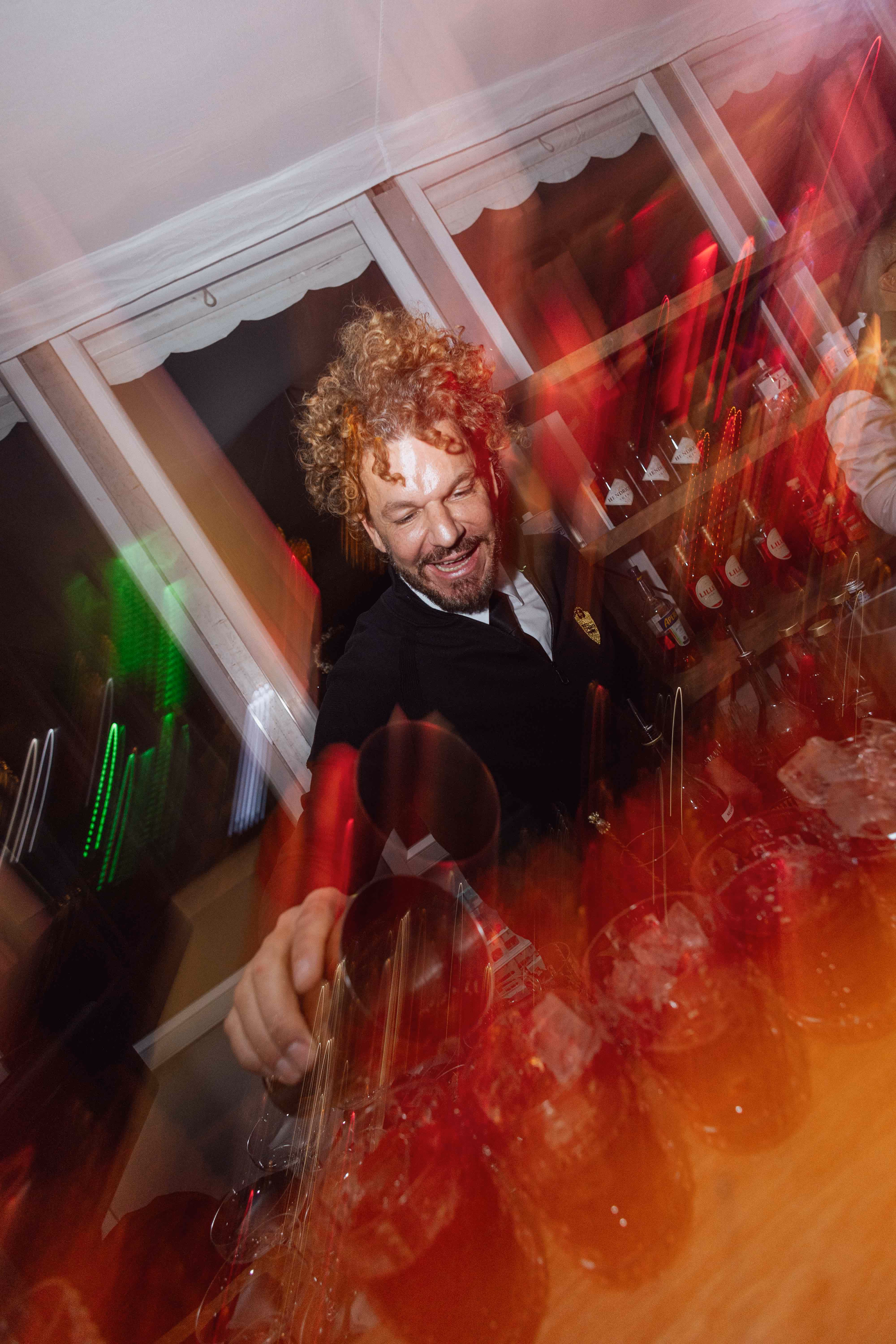 Mario Hofferer with curly hair pouring drinks behind a bar with ice-filled glasses and bottles.
