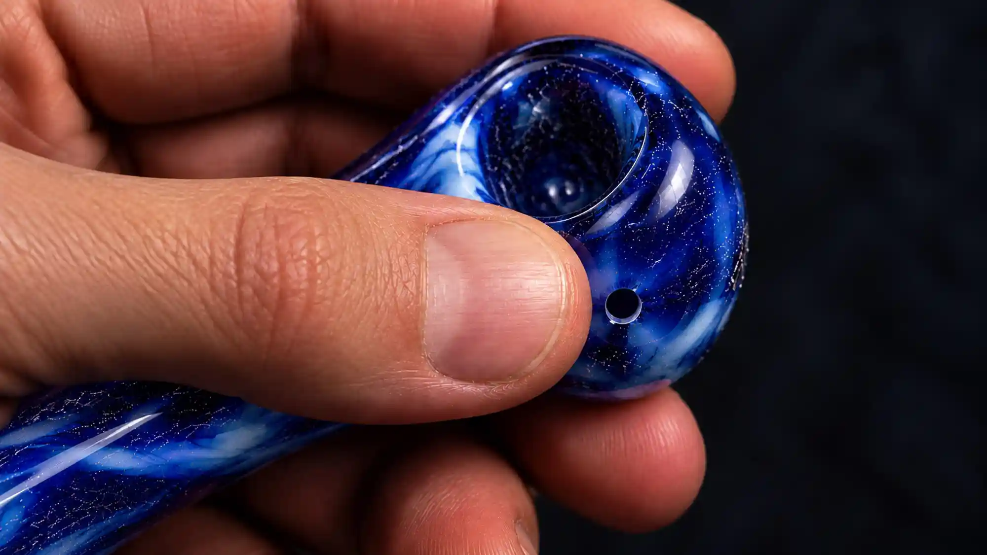 A thumb lifting off the carb hole on the side of a glass spoon pipe