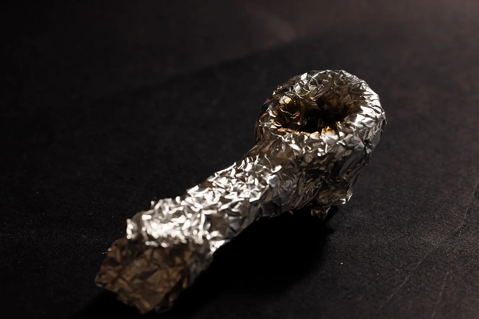 Improvised pipe made from crumpled aluminum foil, with a scorched bowl, on a dark surface