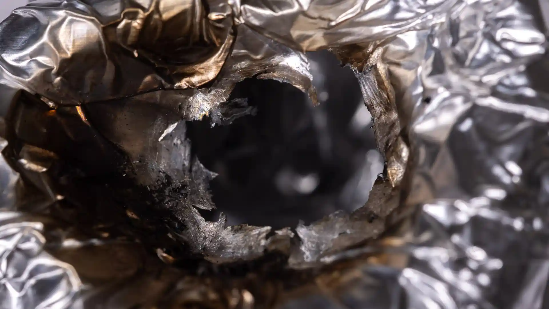 Close-up of a hole where aluminum foil melted through under a lighter flame, with torn edges and heat discoloration