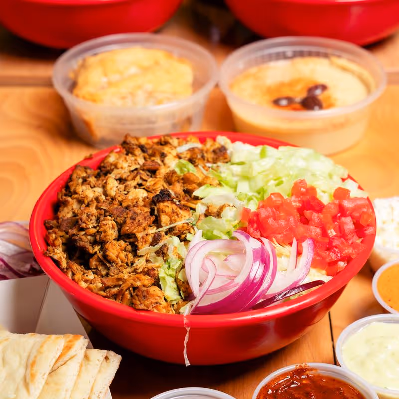 Red bowl filled with grilled chicken, diced tomatoes, shredded lettuce, and sliced red onions, surrounded by small containers of sauces and pita bread.