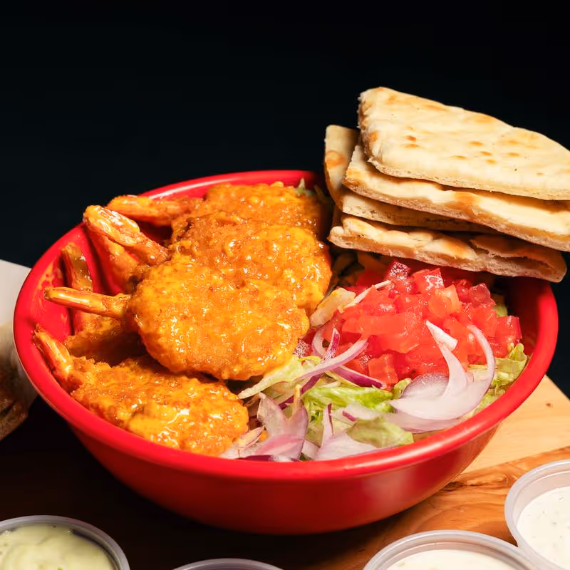 Red bowl with coated shrimp, chopped tomatoes, sliced onions, lettuce, and folded flatbread stacked on top.