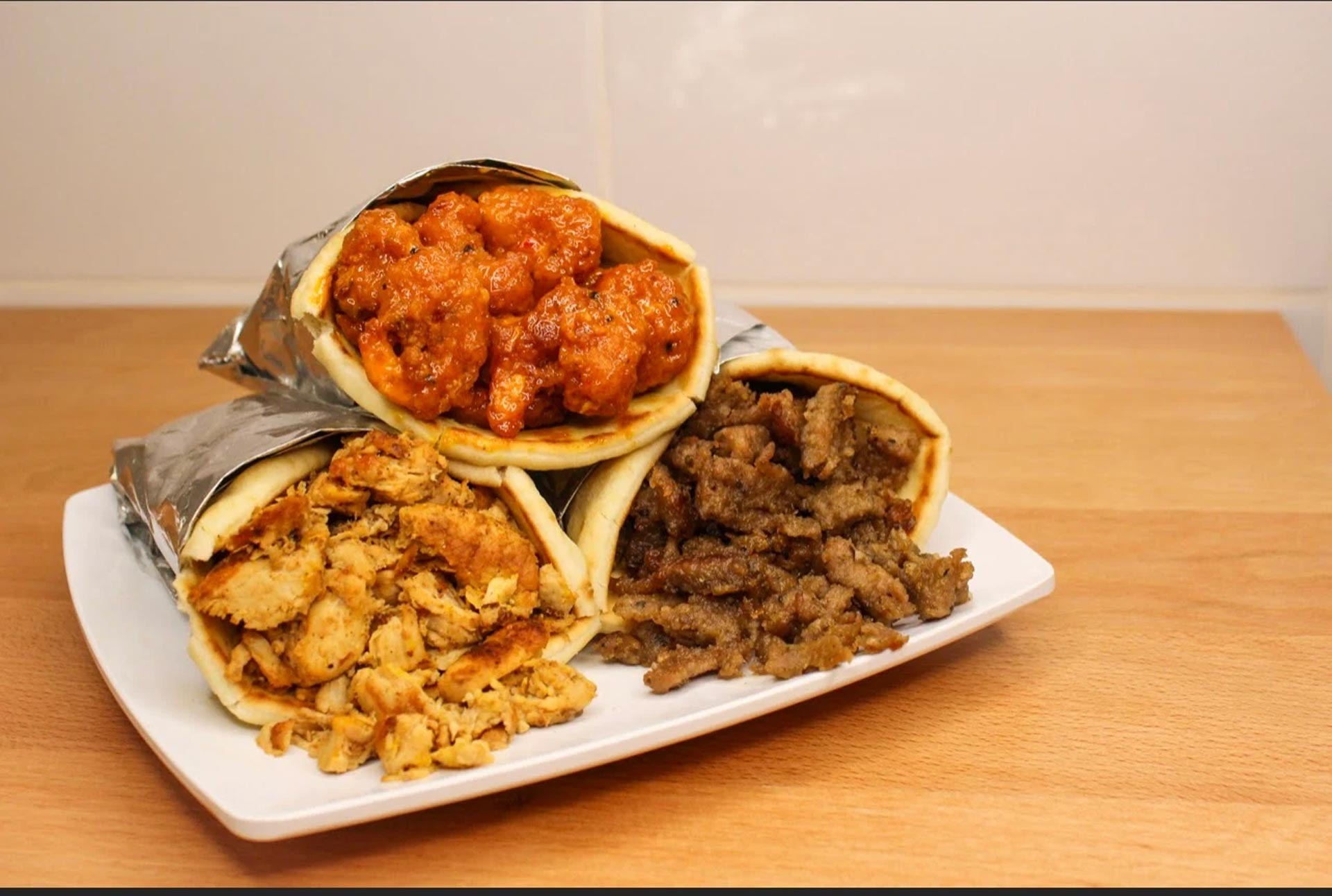 Three pita wraps with different fillings on a white plate on a wooden surface, featuring crispy orange sauce-coated bites, spiced shredded chicken, and grilled beef strips.