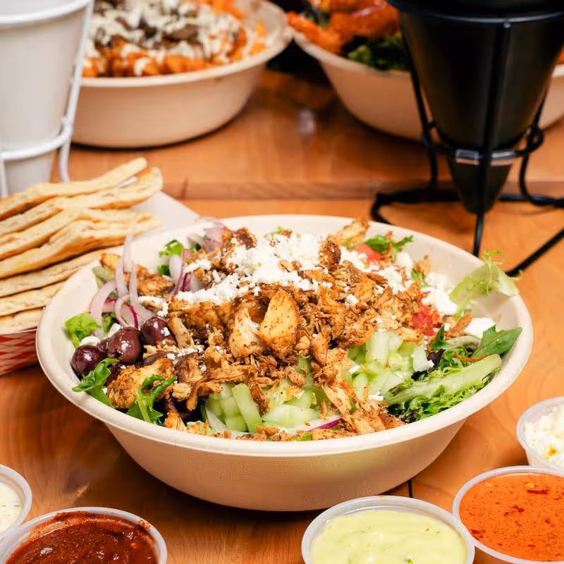 Bowl of Greek salad with grilled chicken, cucumbers, onions, olives, feta cheese, and greens accompanied by pita bread and various dipping sauces.