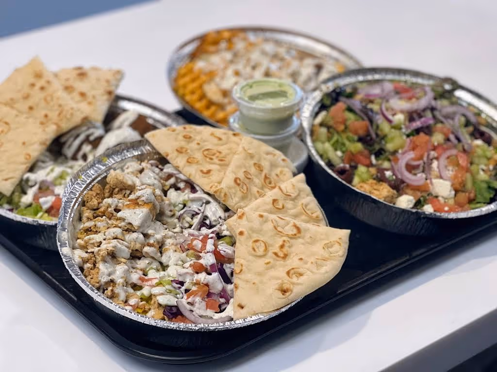 Tray with three foil containers of Mediterranean food including a salad with tomatoes, cucumbers, and onions, a dish with grilled chicken and pita bread, and another dish with a creamy dipping sauce.