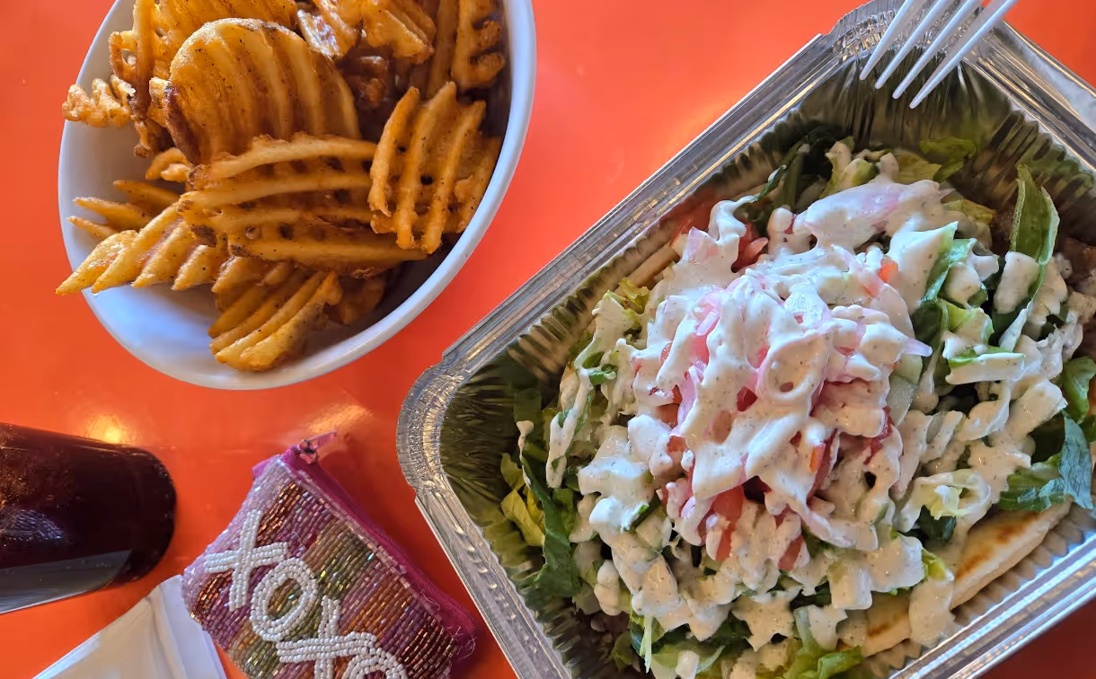 Aluminum container with salad topped with creamy dressing, a bowl of waffle fries, a drink, and a colorful beaded pouch on an orange table.