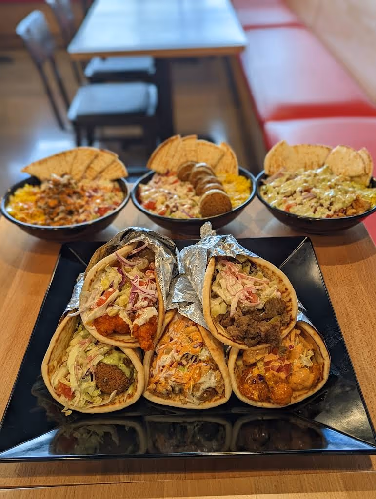 A black plate with five assorted wraps filled with meats, vegetables, and sauces, accompanied by three bowls of salad with pita chips on a wooden table.