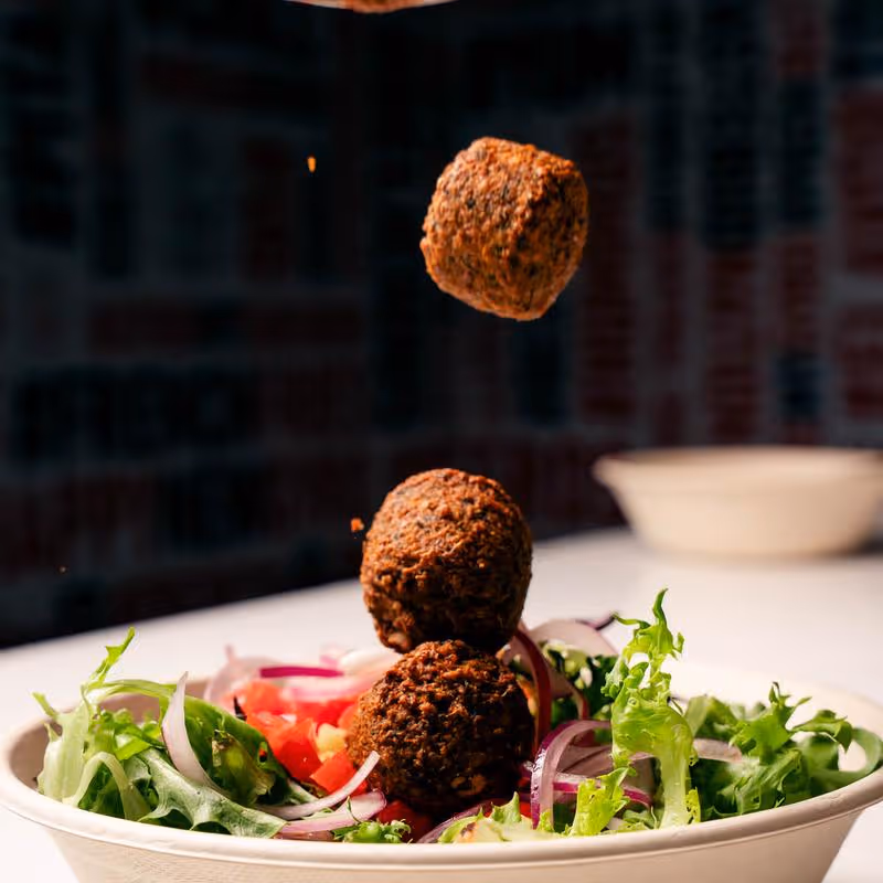 Three falafel balls falling into a bowl filled with fresh green lettuce and sliced red onions.