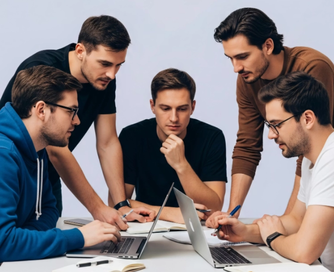 Group of five young men gathered around a table working together on laptops and notebooks.