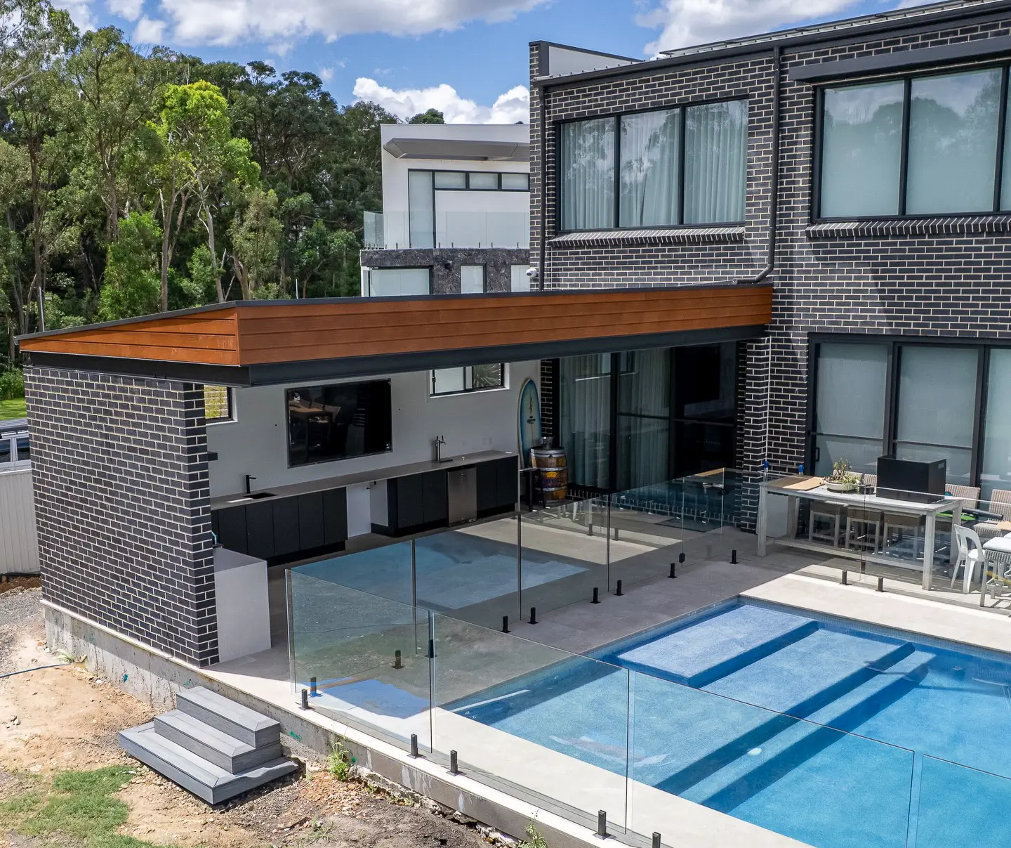 Modern house patio with glass fence surrounding a blue swimming pool and an outdoor kitchen under a wooden roof.