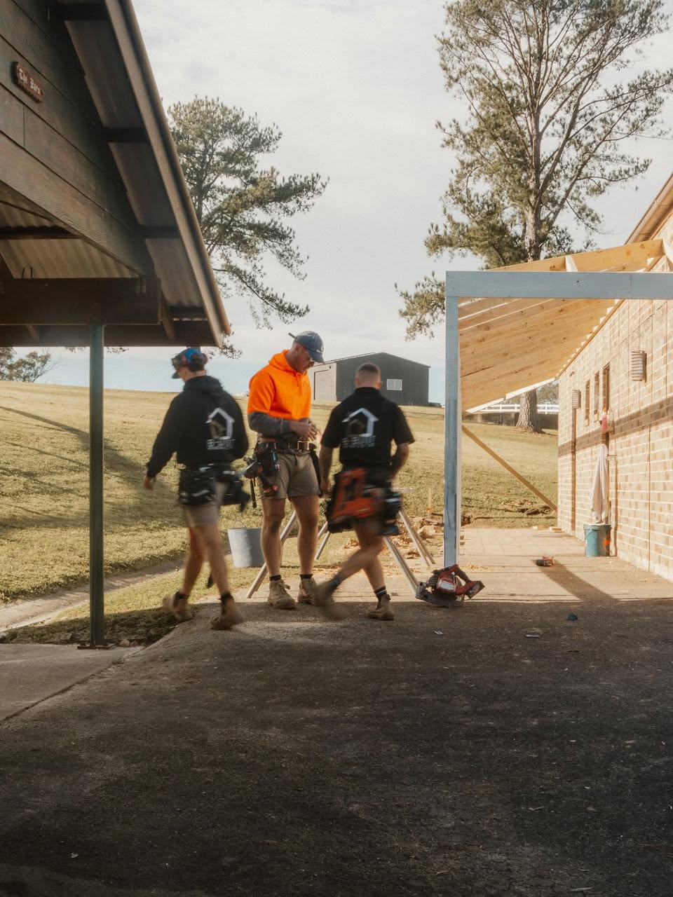 Three construction workers standing and walking near a partially built wooden frame attached to a brick building.
