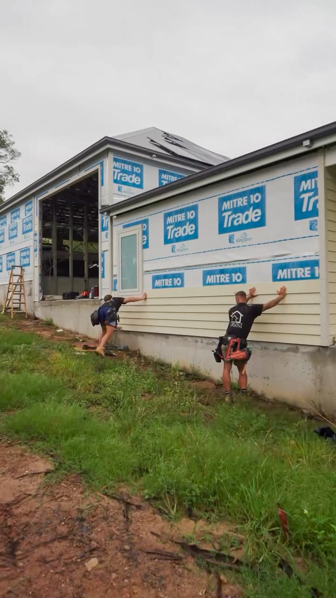 Two construction workers installing beige siding on a building wrapped with Mitre 10 Trade branded house wrap.