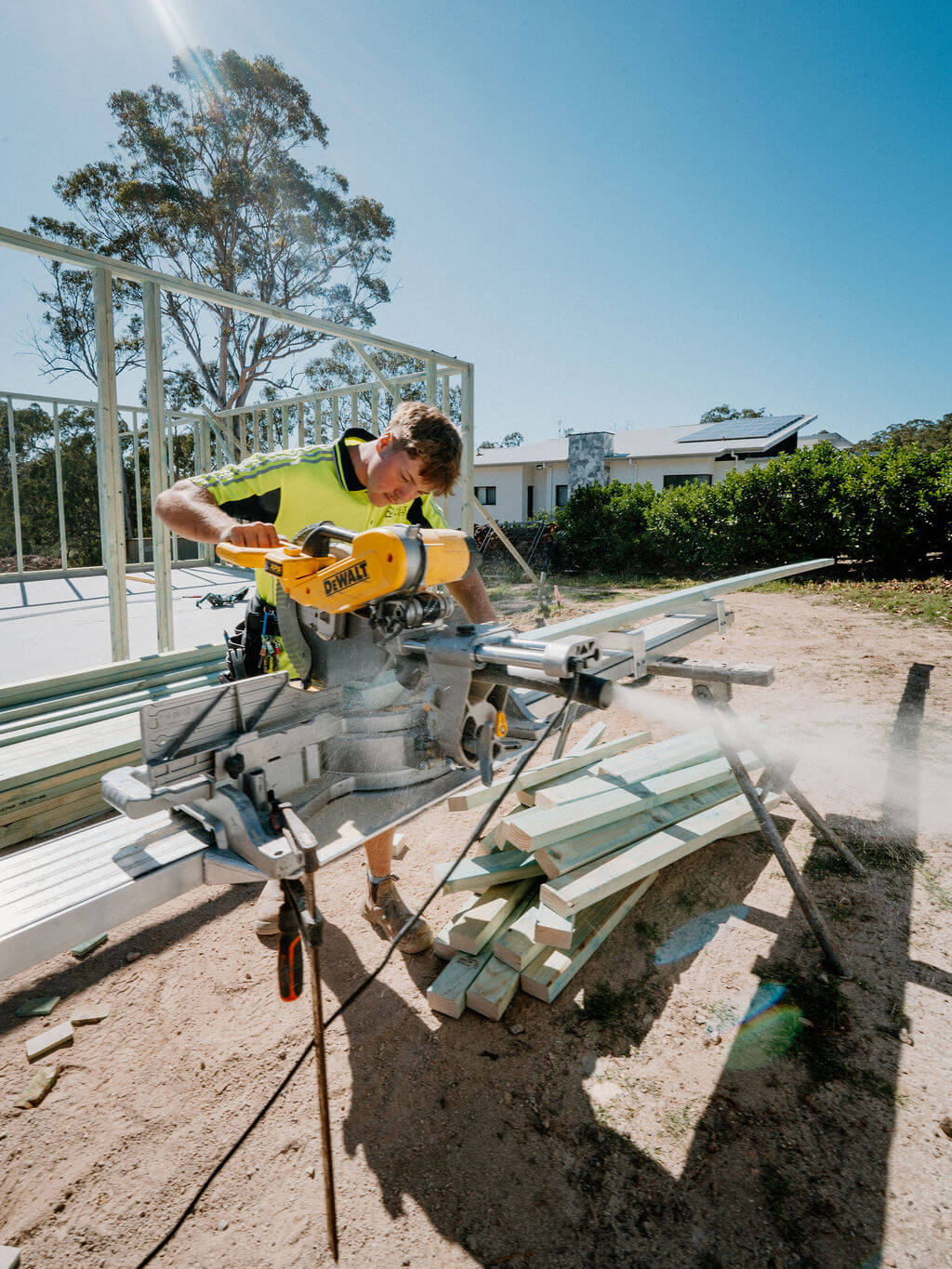 Construction worker in bright yellow shirt operating a DeWalt miter saw to cut wooden planks at an outdoor building site.