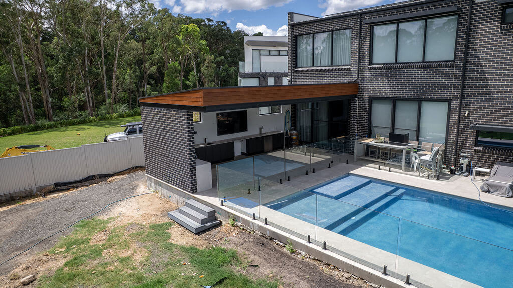 Modern backyard with a glass-fenced swimming pool, patio with outdoor kitchen, and a two-story brick house.