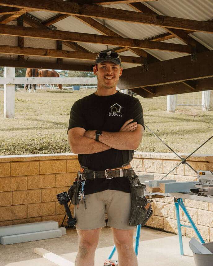 Man in a B Built Projects shirt and tool belt standing confidently in a wooden framed outdoor workspace.