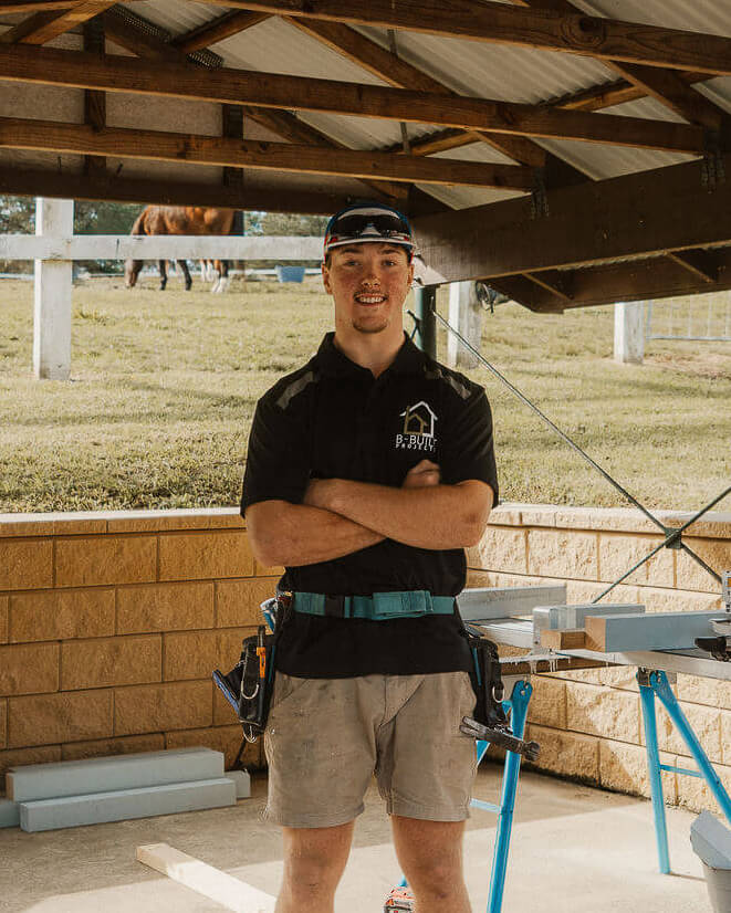 A young man in a black B-Built polo shirt and khaki shorts stands with arms crossed in a workshop with woodworking tools and horses visible outside.