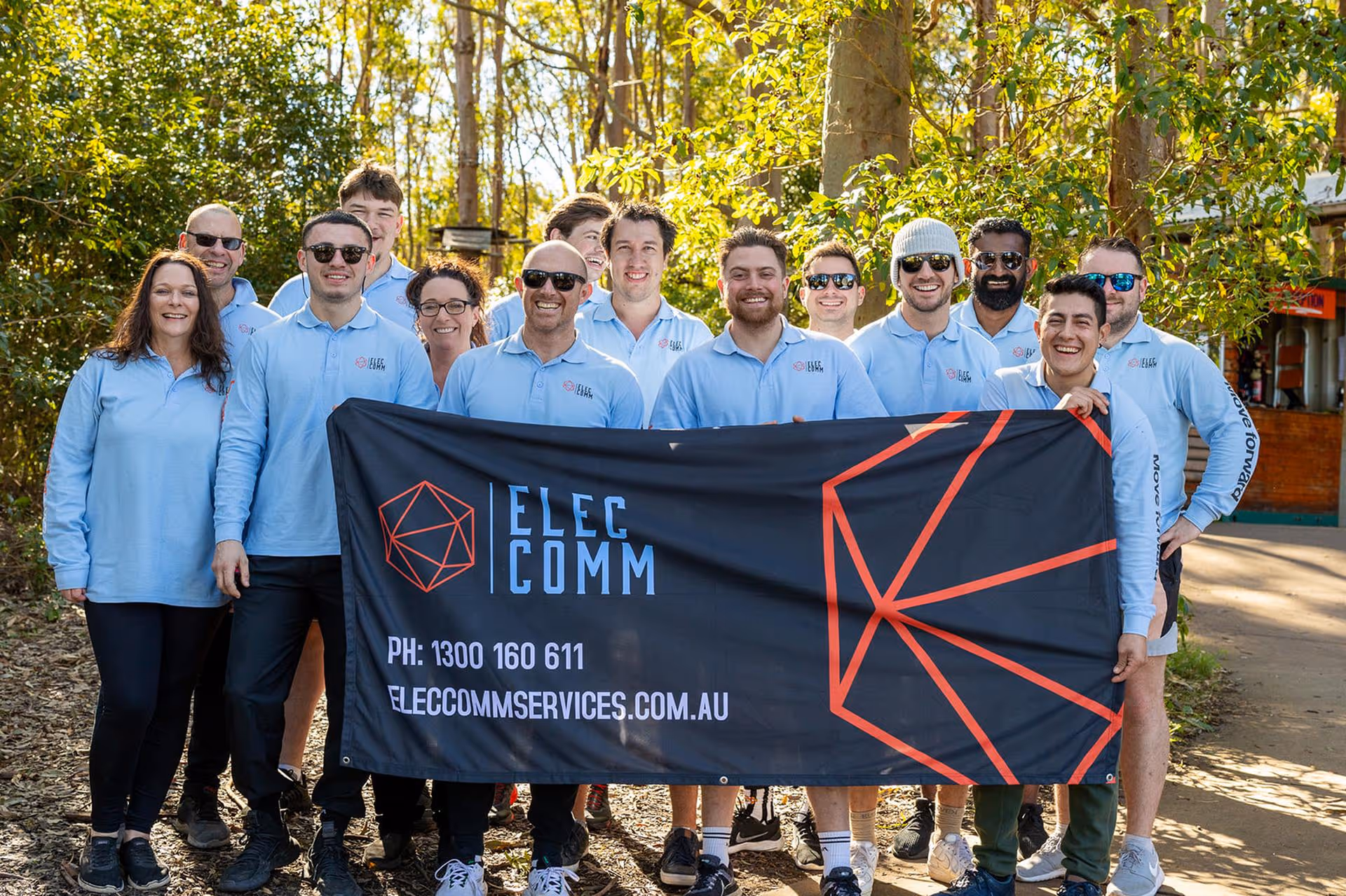 Group of smiling people wearing light blue Elec Comm shirts holding a black Elec Comm banner outdoors with trees in the background.