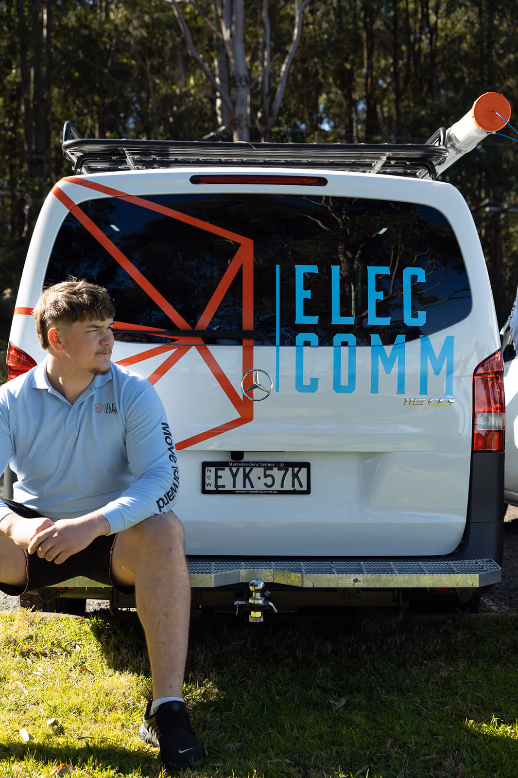 Man in a light blue ElecComm shirt sitting on the back bumper of a white ElecComm van with a forest background.