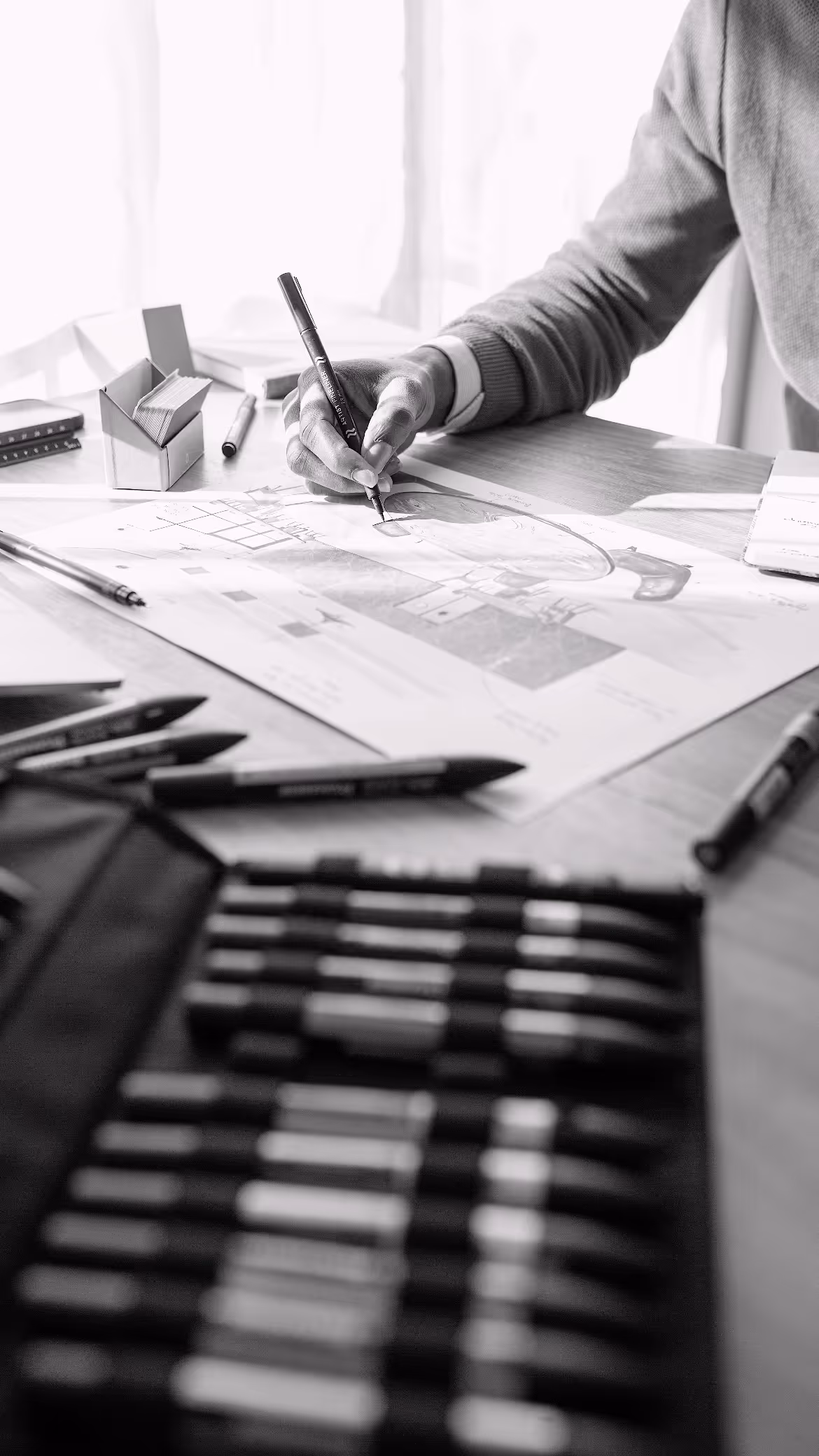 Personne dessinant une illustration détaillée avec un marqueur sur une table entourée de crayons et stylos.