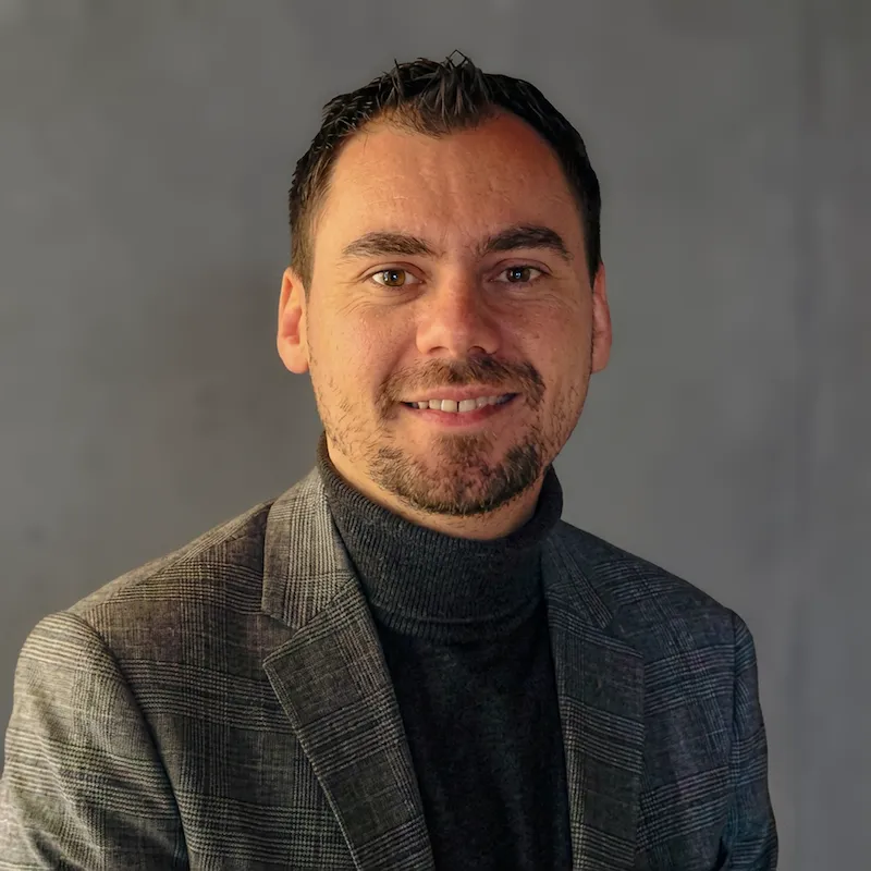 Portrait d'un homme souriant portant un col roulé noir et une veste grise à carreaux devant un mur gris flou.