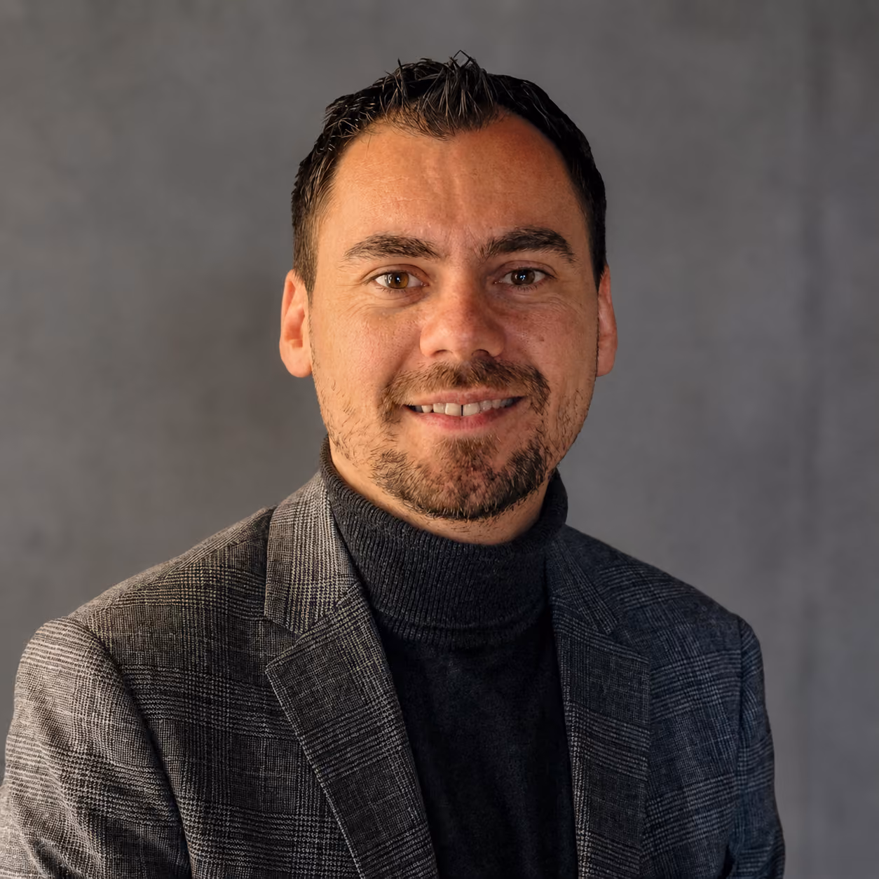 Portrait d'un homme souriant portant un col roulé noir et une veste grise à carreaux devant un mur gris flou.