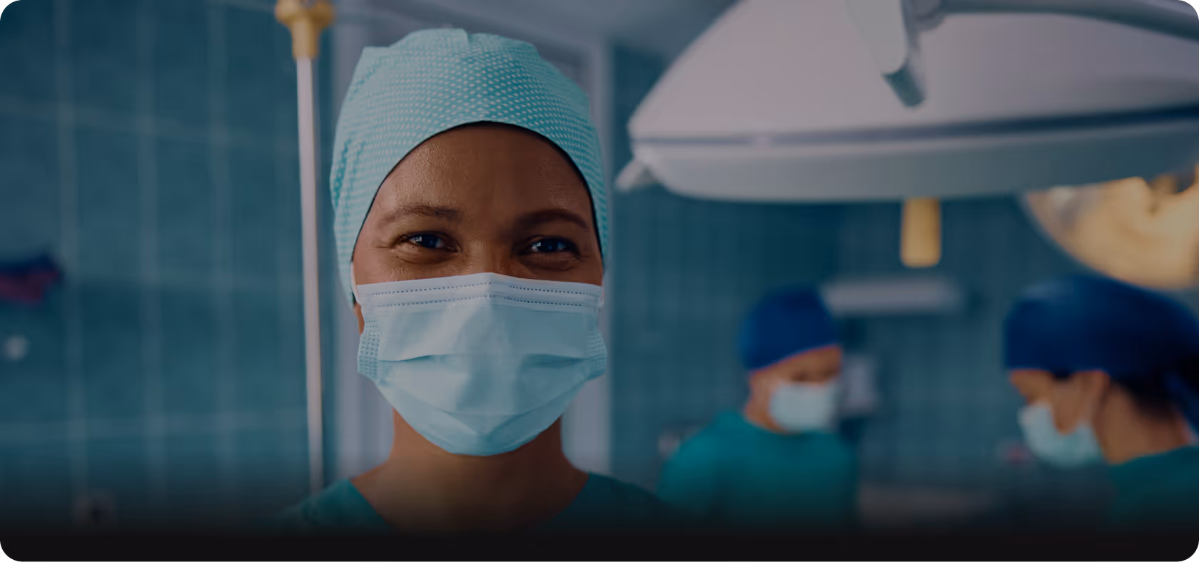 Smiling healthcare professional wearing surgical scrubs, cap, and mask in an operating room with two medical staff blurred in the background.