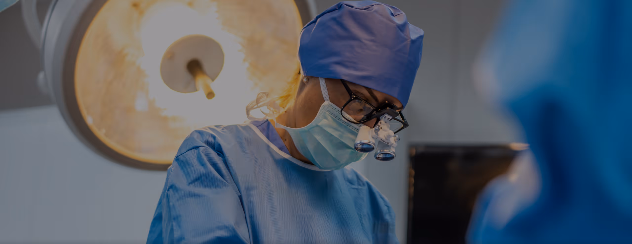 Surgeon wearing magnifying glasses and surgical mask under bright operating room light.