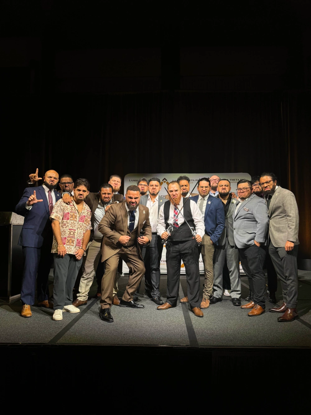 Group of men dressed in formal and semi-formal attire posing with serious expressions on a stage with a Lambda Theta Pi banner in the background.